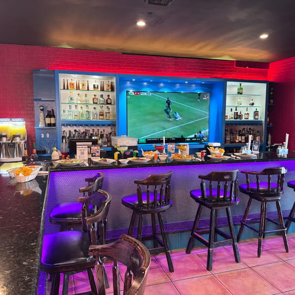A vibrant bar with a large TV showing soccer, colorful drinks, and bar stools under purple lighting.