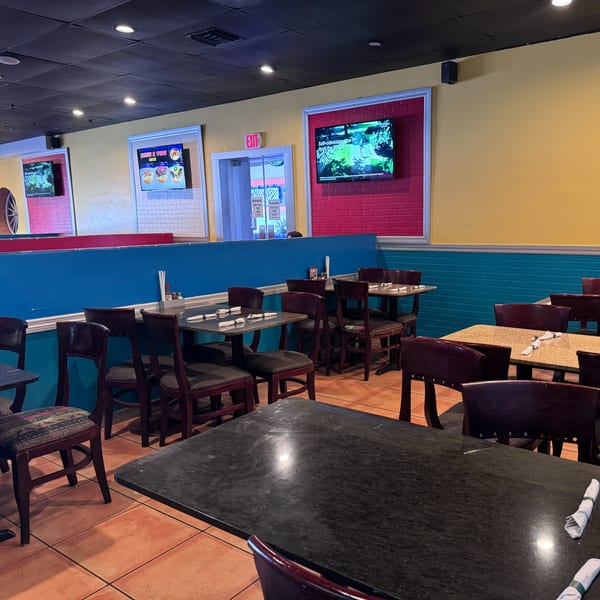 Empty restaurant dining room with colorful walls, dark tables, and chairs, featuring TVs and an exit sign.